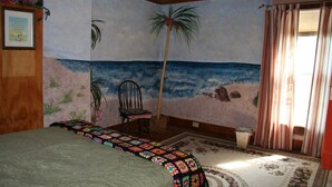 Een slaapkamer met een strandmuur, een bed met een kleurrijke deken, een stoel en een raam met gordijnen.