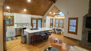 A spacious living area with a high ceiling and wooden beams, a stone fireplace, and a kitchen with white cabinets and a central island. - Lake house with boat ramp access across the street & tons of boat parking space. (Monticello)