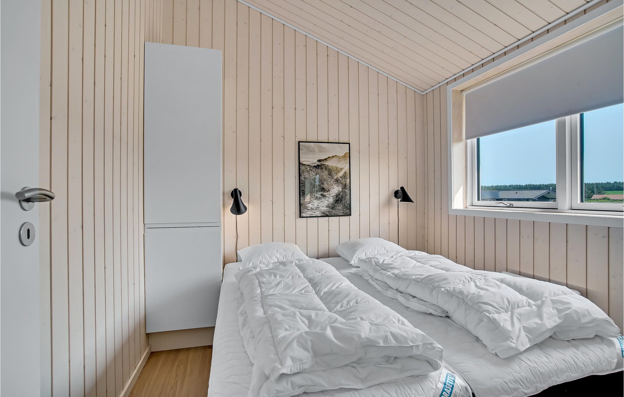A bedroom with a bed, a wardrobe, a window with a view, and a wall-mounted lamp.