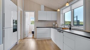 A modern kitchen with white cabinetry, a wooden floor, and a view of the countryside through large windows. - Lovely home in Løkken with sauna (Løkken)