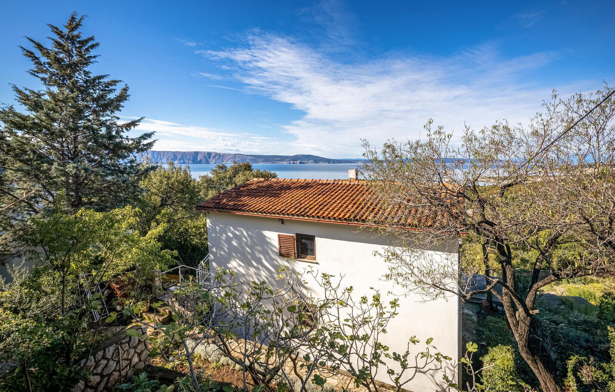 A white building with a tiled roof, surrounded by trees and vegetation, overlooking a body of water and distant mountains.