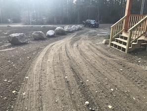Leading to a secluded cabin, a dirt road winds through the forest, flanked by large rocks and a wooden staircase with a handrail.