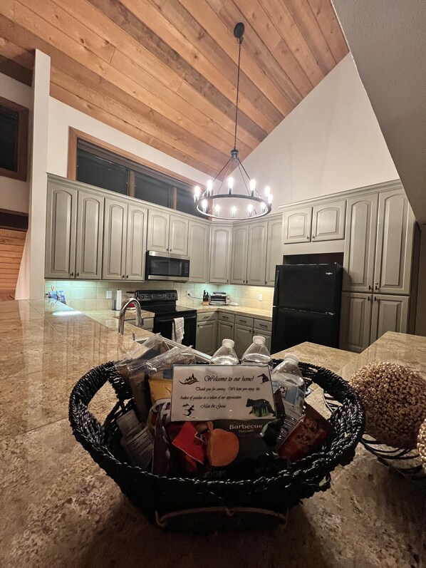 A kitchen with light gray cabinets, a black refrigerator, and a chandelier. There is a basket with a welcome sign and snacks on the counter. - Beaver Creek Retreat! Spacious 3BR+Loft Ski In/Out – Steps to Adventure (Beaver Creek)