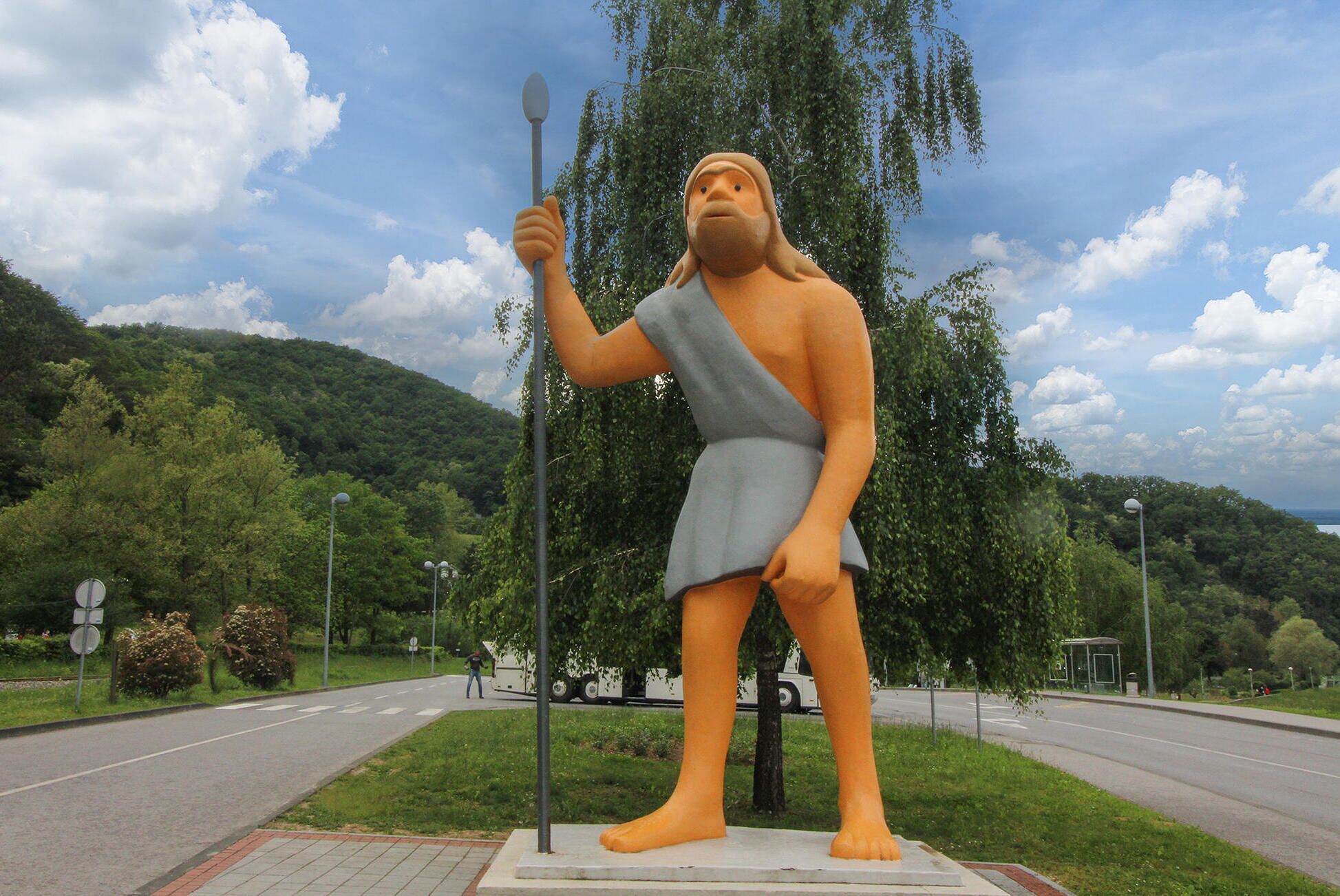 Una estatua de una figura sosteniendo una lanza, con un camino, árboles y un paisaje montañoso de fondo.