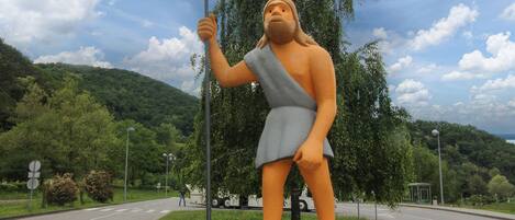 Una estatua de una figura sosteniendo una lanza, con un camino, árboles y un paisaje montañoso de fondo.