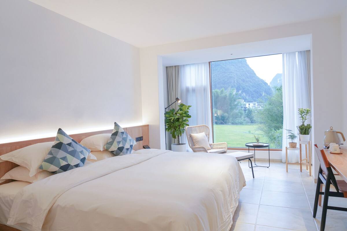 A hotel room with a large bed, a chair, a small table, and a view of a mountainous landscape through a large window.
