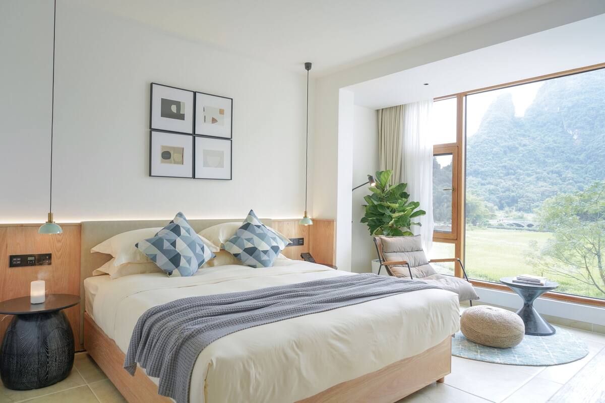 A modern bedroom with a large bed, a bedside table, a nightstand, a potted plant, and a view of the outdoors.