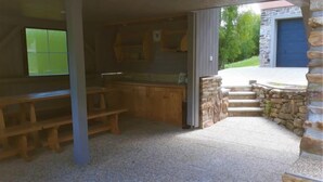 A covered outdoor kitchen area with wooden benches, a stone wall, and a view of a garage. - Nebahovy u Prachatic by Interhome (Prachatice)