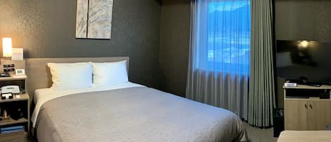 A hotel room with a large bed, a flat-screen TV, a bedside table, a lamp, and a window with curtains.