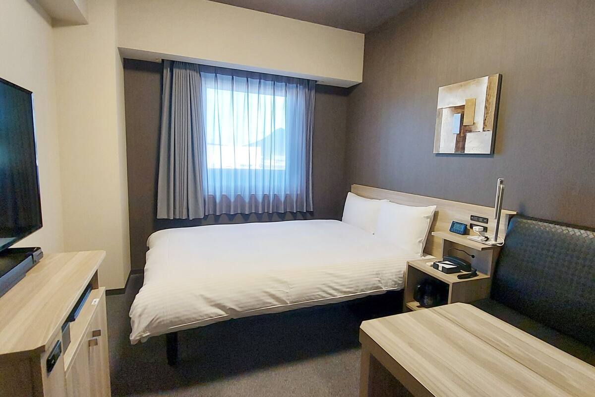 A hotel room with a bed, a desk, a chair, a TV, a bedside table, and a window with curtains.