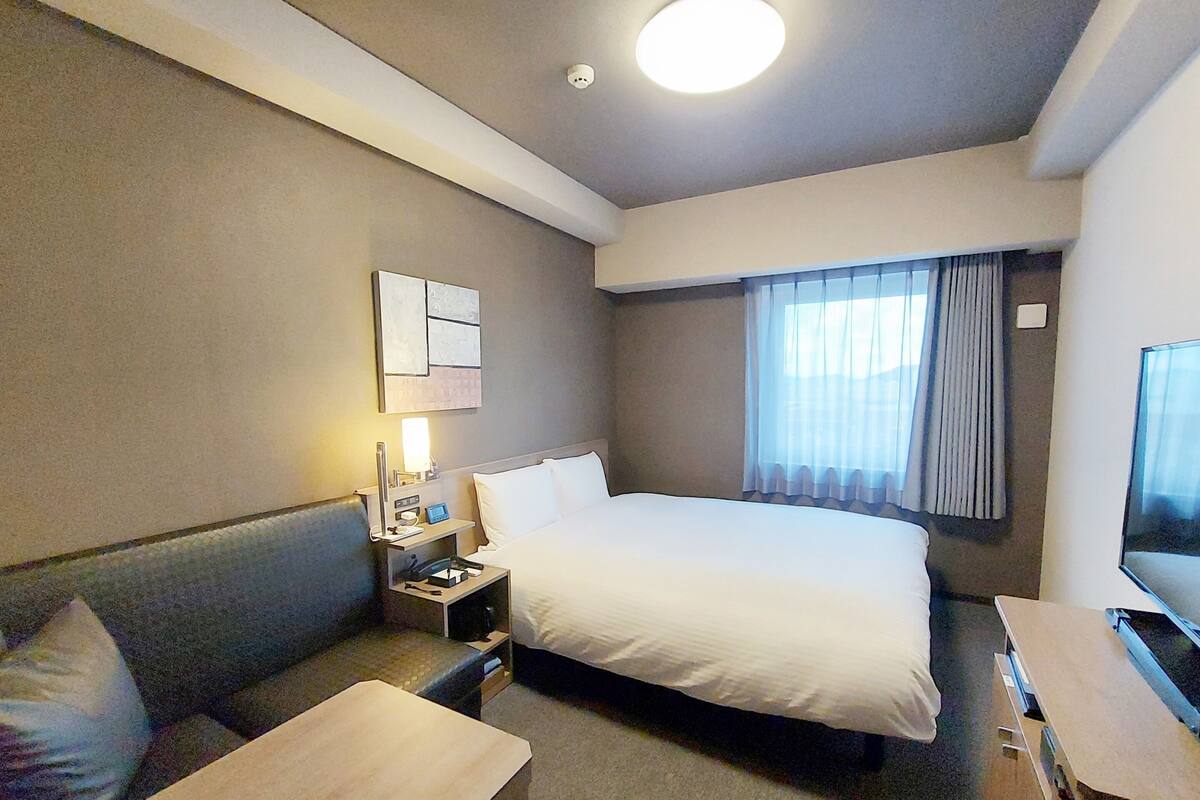 A hotel room with a bed, a desk, a chair, a nightstand, a TV, and a window with curtains.