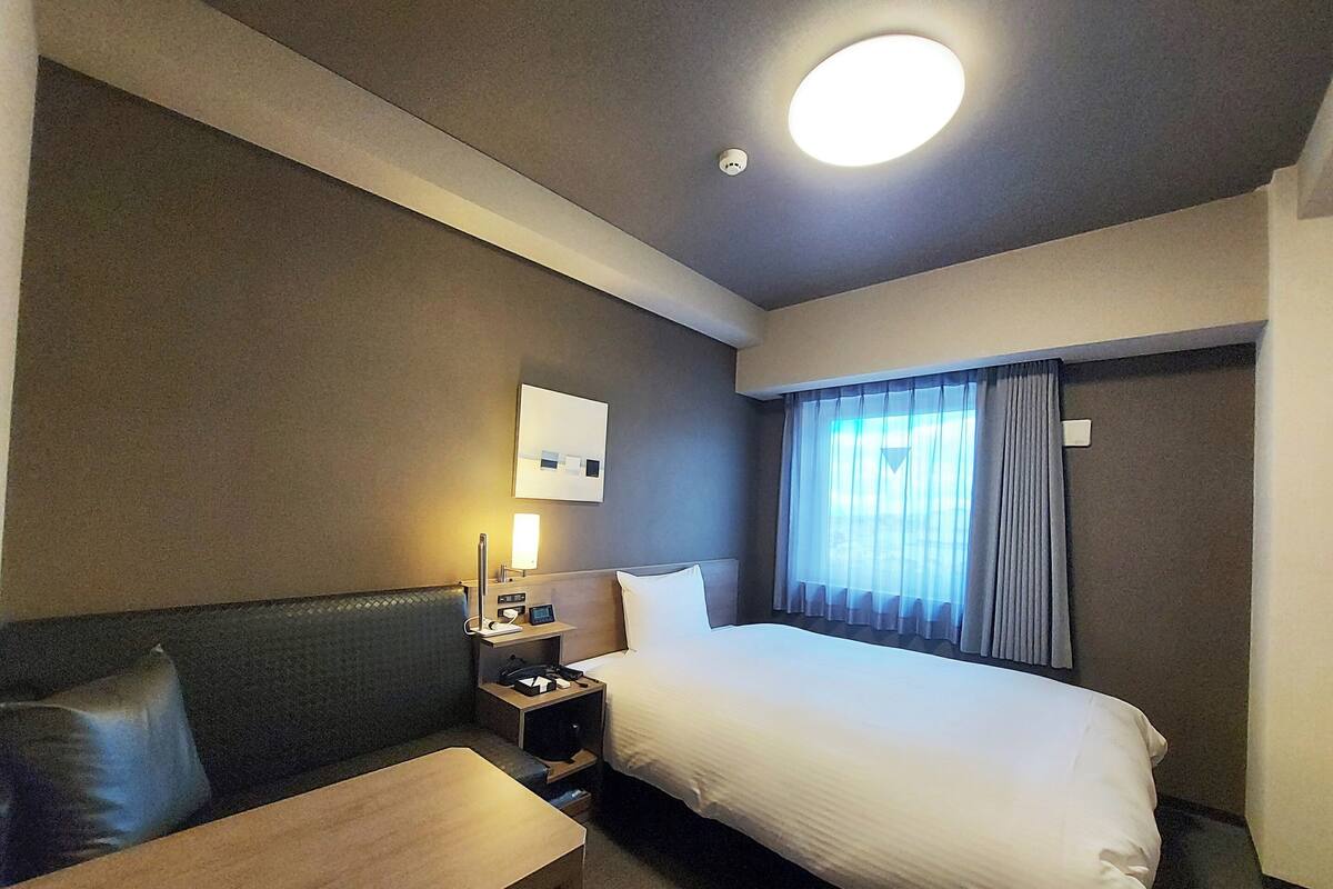 A hotel room with a bed, a desk, a chair, a nightstand, a lamp, and a window with curtains.