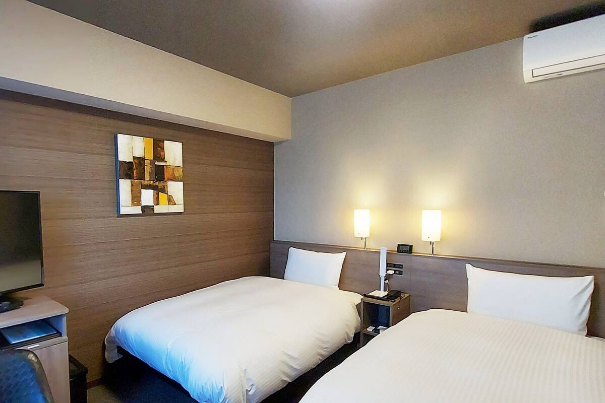 A hotel room with two single beds, a desk with a chair, a flat-screen TV, and a wall-mounted air conditioner.
