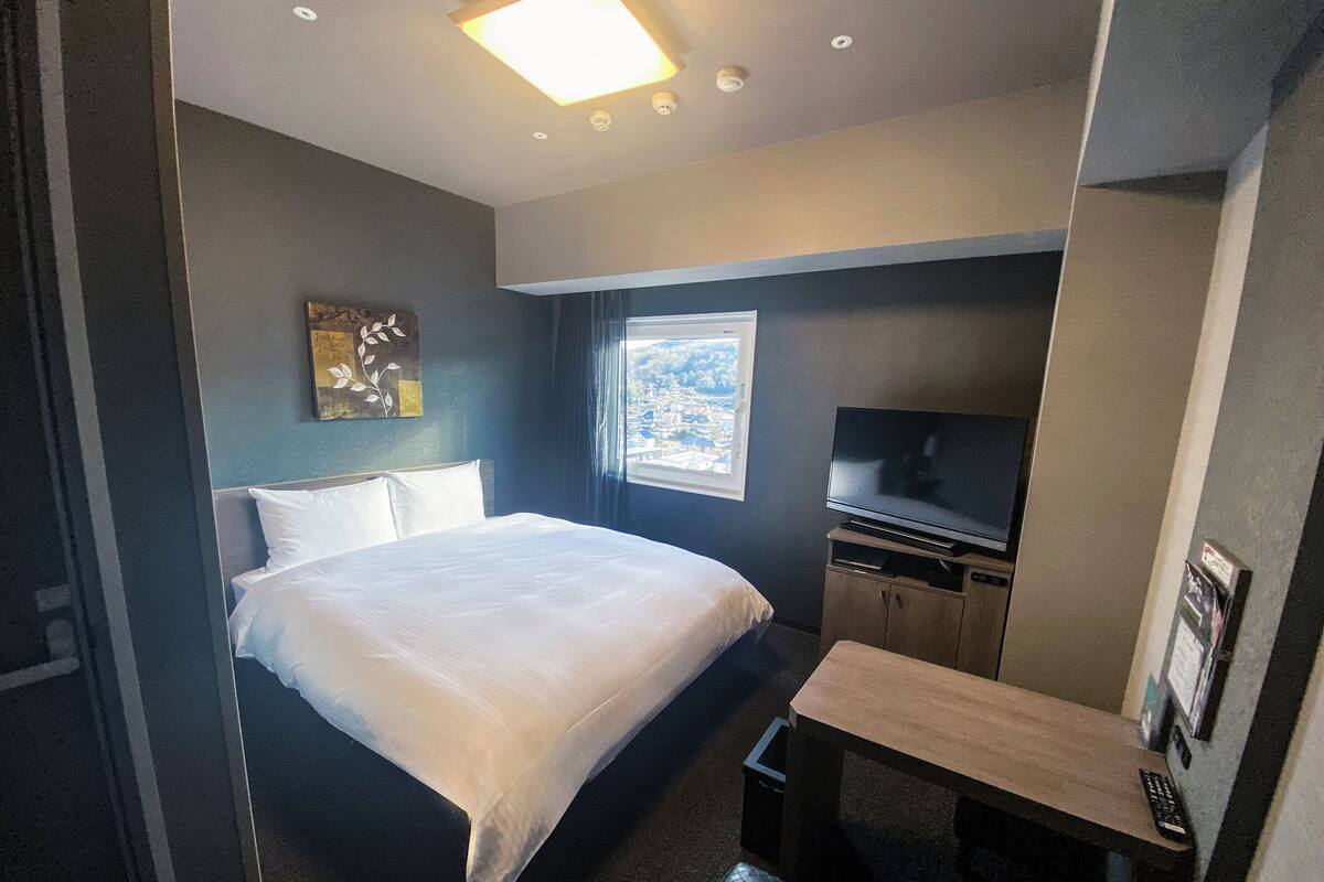 Ett hotellrum med en säng, en tv, ett litet bord och ett fönster med utsikt över staden.