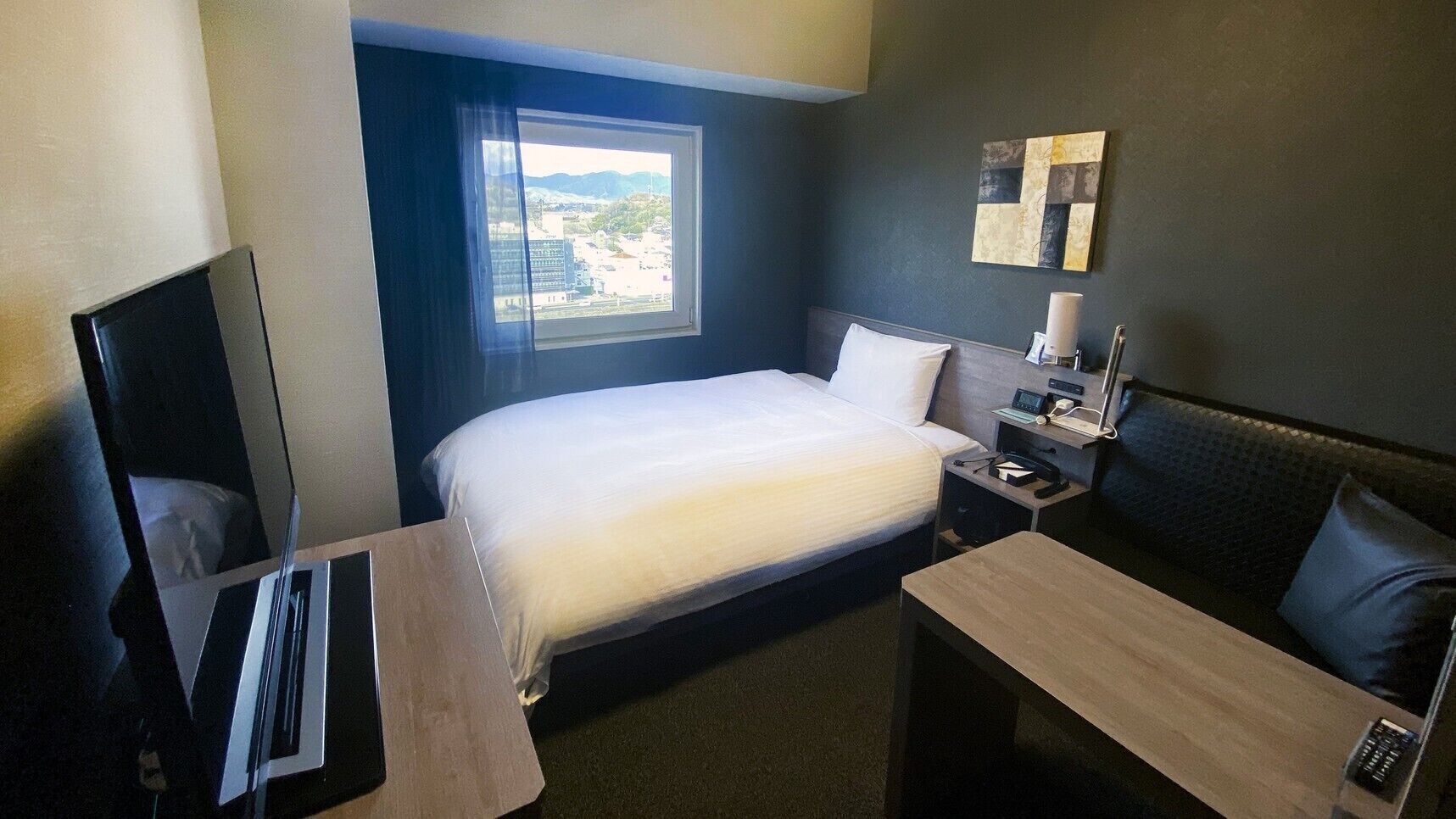 A hotel room with a bed, a desk, a chair, a TV, a bedside table with a lamp, and a window with a view of buildings.