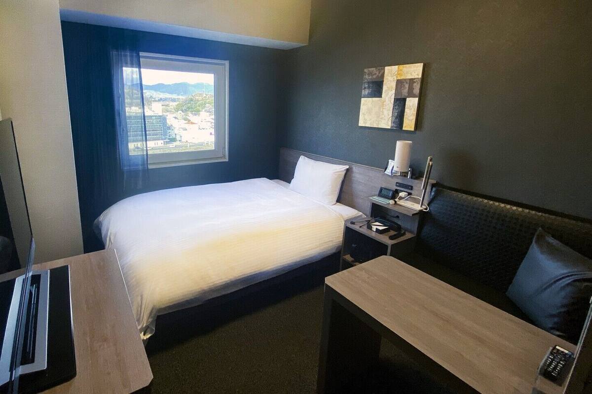 Ett hotellrum med en säng, ett skrivbord, en stol, en TV, ett nattduksbord med en lampa och ett fönster med utsikt över byggnader.