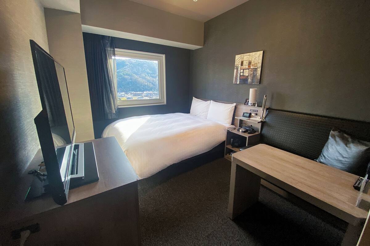 Ett hotellrum med en säng, ett skrivbord, en stol, ett nattduksbord, en lampa och ett fönster med utsikt över bergen.
