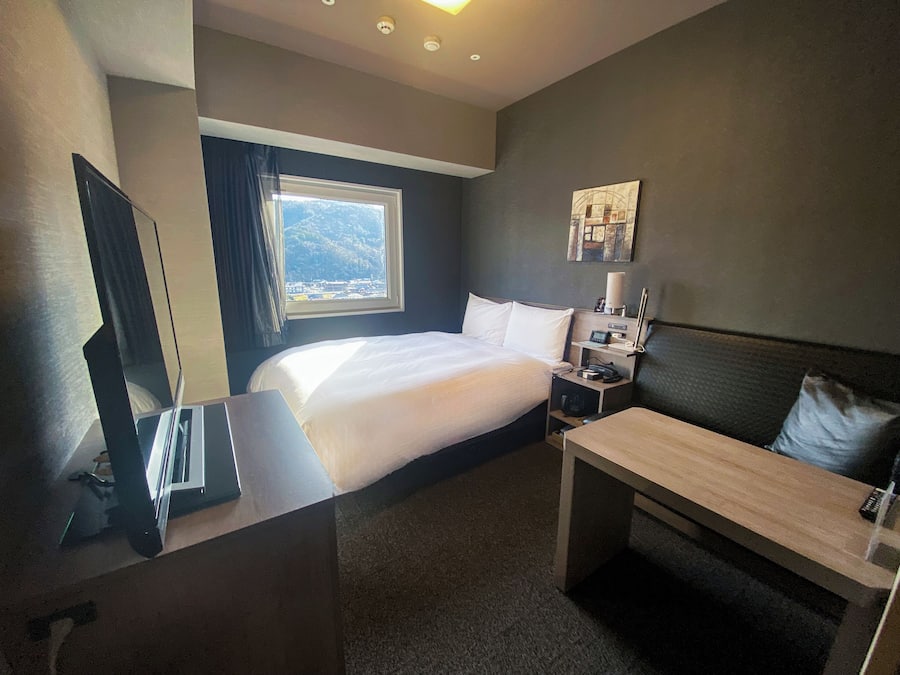 A hotel room with a bed, a desk, a chair, a nightstand, a lamp, and a window with a view of mountains.
