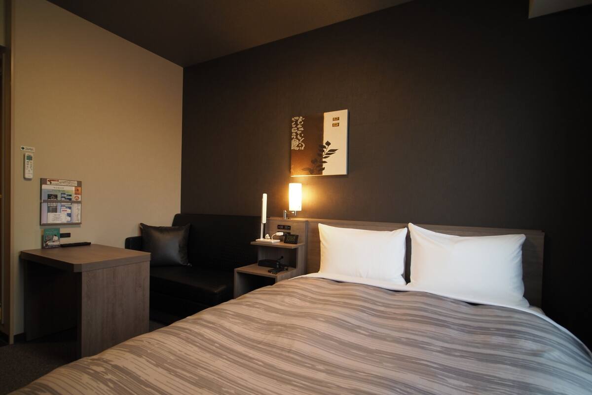 A hotel room with a bed, a desk, a chair, and a nightstand with a lamp.