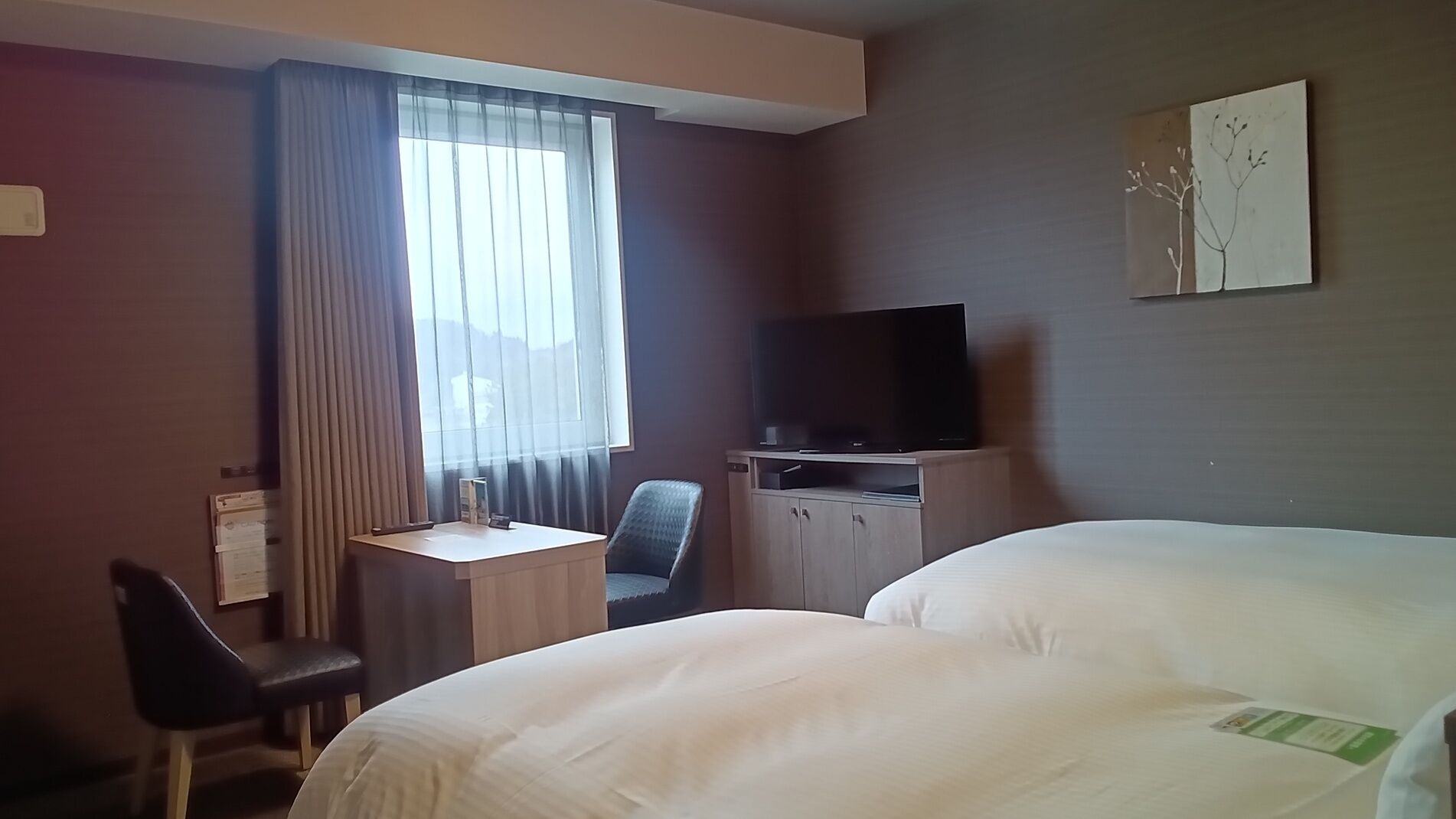 Une chambre d’hôtel avec un lit, un bureau et une chaise, une télévision et une fenêtre avec des rideaux.