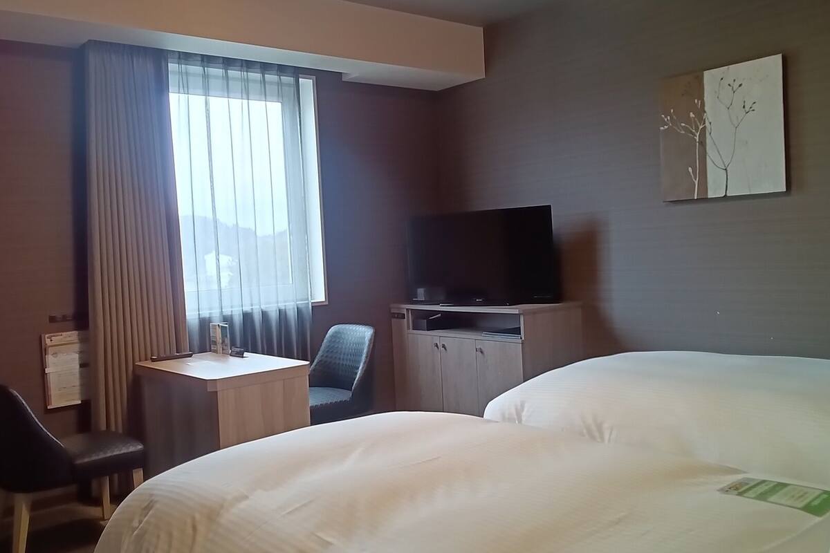 A hotel room with a bed, a desk with a chair, a television, and a window with curtains.