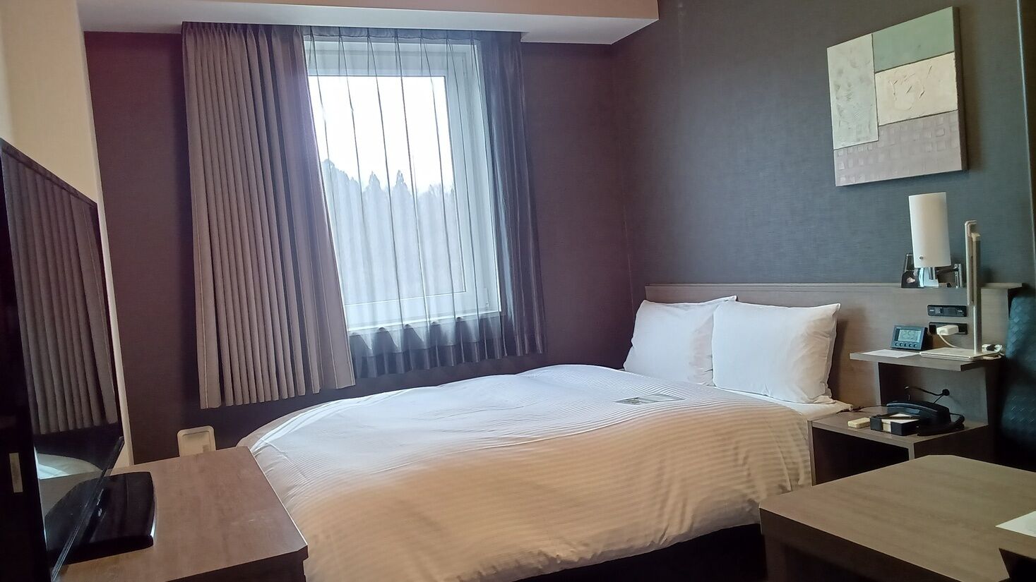 Une chambre d’hôtel avec un lit, un bureau, une chaise, une table de chevet, une lampe et une fenêtre avec des rideaux.