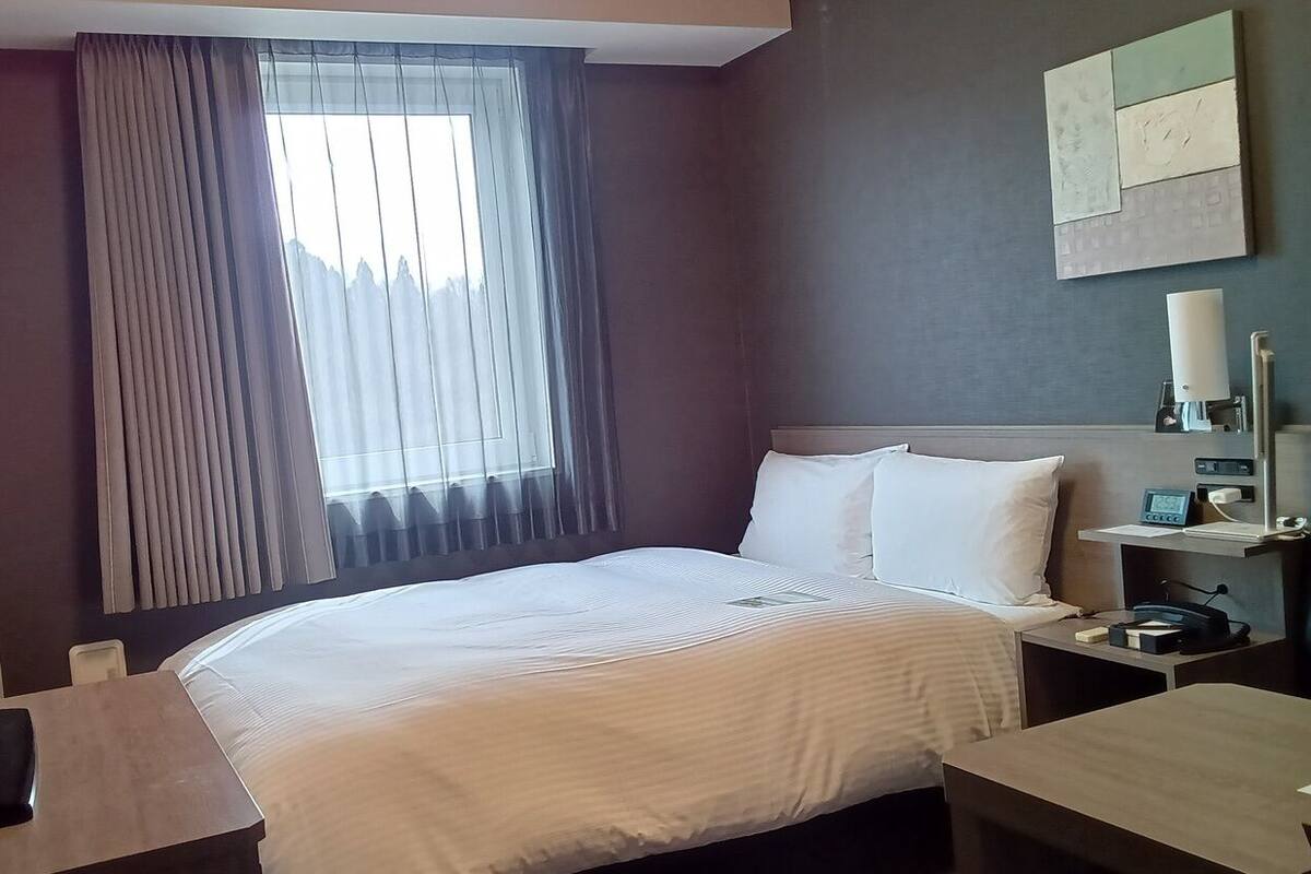 A hotel room with a bed, a desk, a chair, a nightstand, a lamp, and a window with curtains.