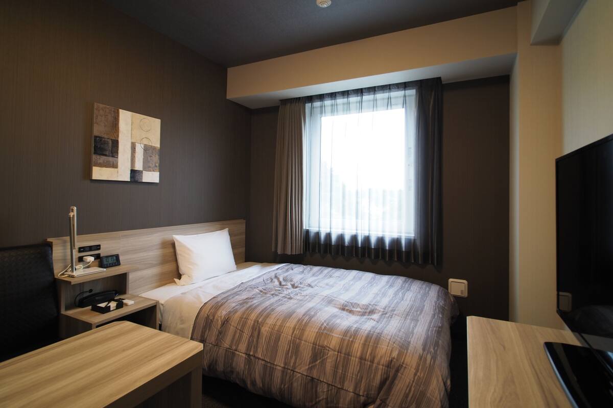 A hotel room with a bed, a desk, a TV, a nightstand, and a window with curtains.