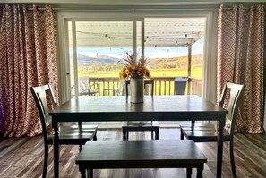 A dining area with a table and chairs, a vase with flowers, and a view of a landscape through large windows.