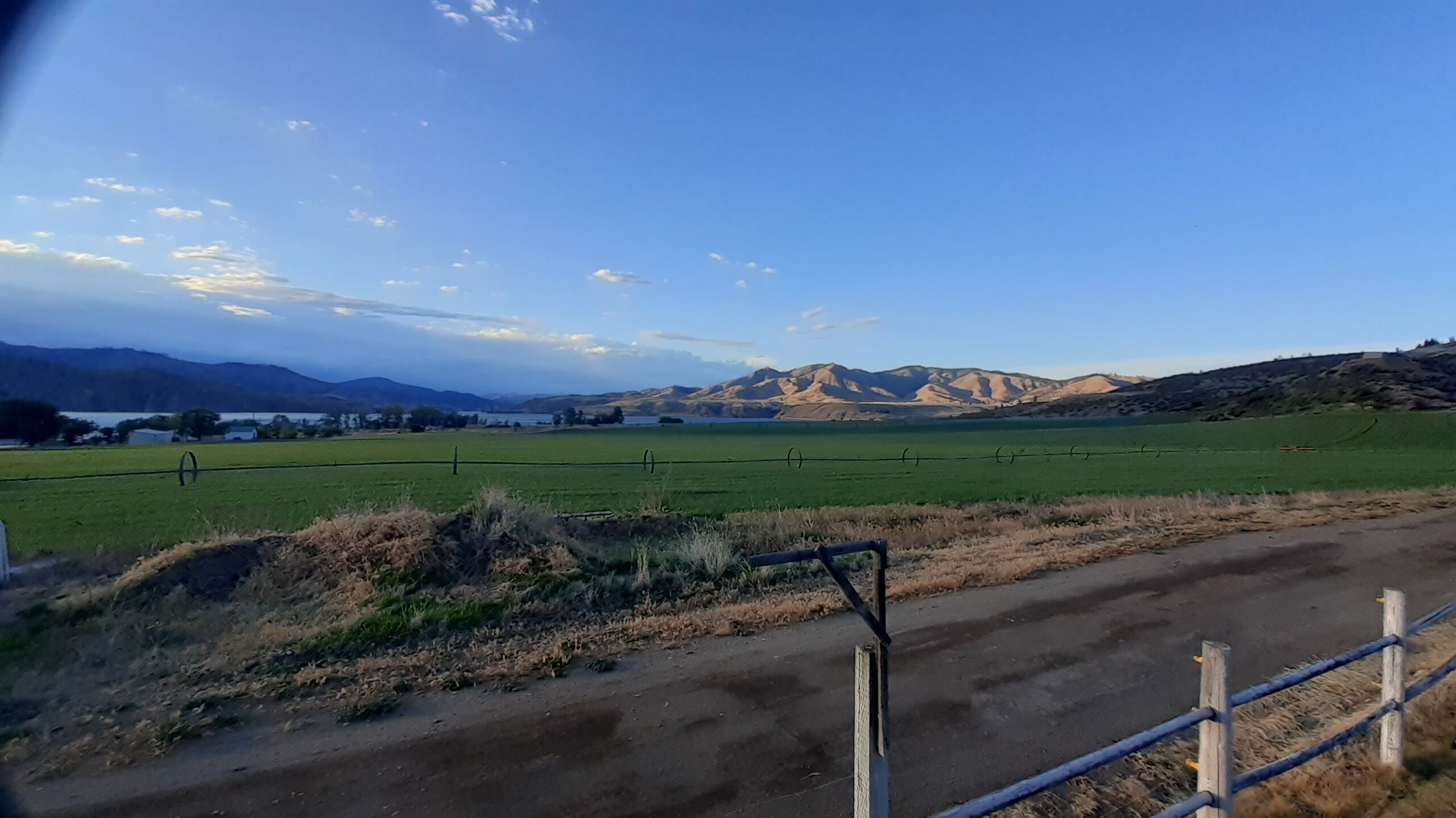 A rural landscape with a dirt road, green fields, and distant mountains under a clear sky.