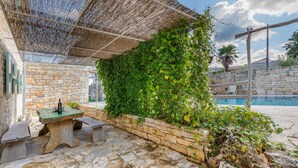 Une terrasse en pierre avec une table et des bancs, une piscine et un mur recouvert de plantes grimpantes.