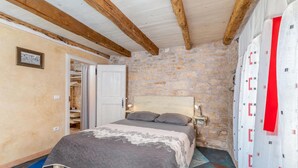 Une chambre avec un mur en pierre, un plafond en bois, un lit recouvert d’une couverture grise, une table de chevet avec une lampe et une armoire.