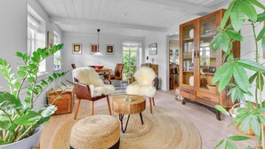 A spacious living room with a high ceiling, wooden flooring, and a large wooden cabinet with glass doors. There are two fluffy chairs, a round wooden coffee table, and several potted plants.