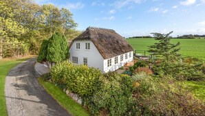 A white cottage with a thatched roof, surrounded by lush greenery and a clear blue sky.