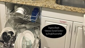 A kitchen with granite countertops, a sink, and a dishwasher. There is a plate of fruit on the counter and a can of soda. The kitchen has white cabinets and a ceiling fan.