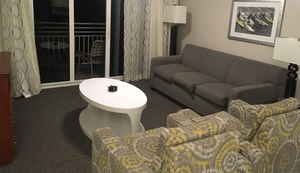 Una sala de estar con un sofá gris, sillones estampados, una mesa de centro blanca y un ventanal corredizo que da a un área exterior.