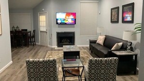 A modern living room with a fireplace, a flat-screen TV mounted above it, a sofa, two patterned armchairs, a coffee table, and a dining area with a table and chairs. - Austin Home (Austin)