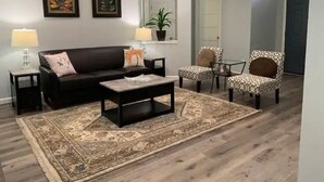 A living room with a dark-colored sofa, two patterned armchairs, a coffee table, and a large rug. There are two wall-mounted lamps, a potted plant, and two framed artworks on the wall. - Austin Home (Austin)