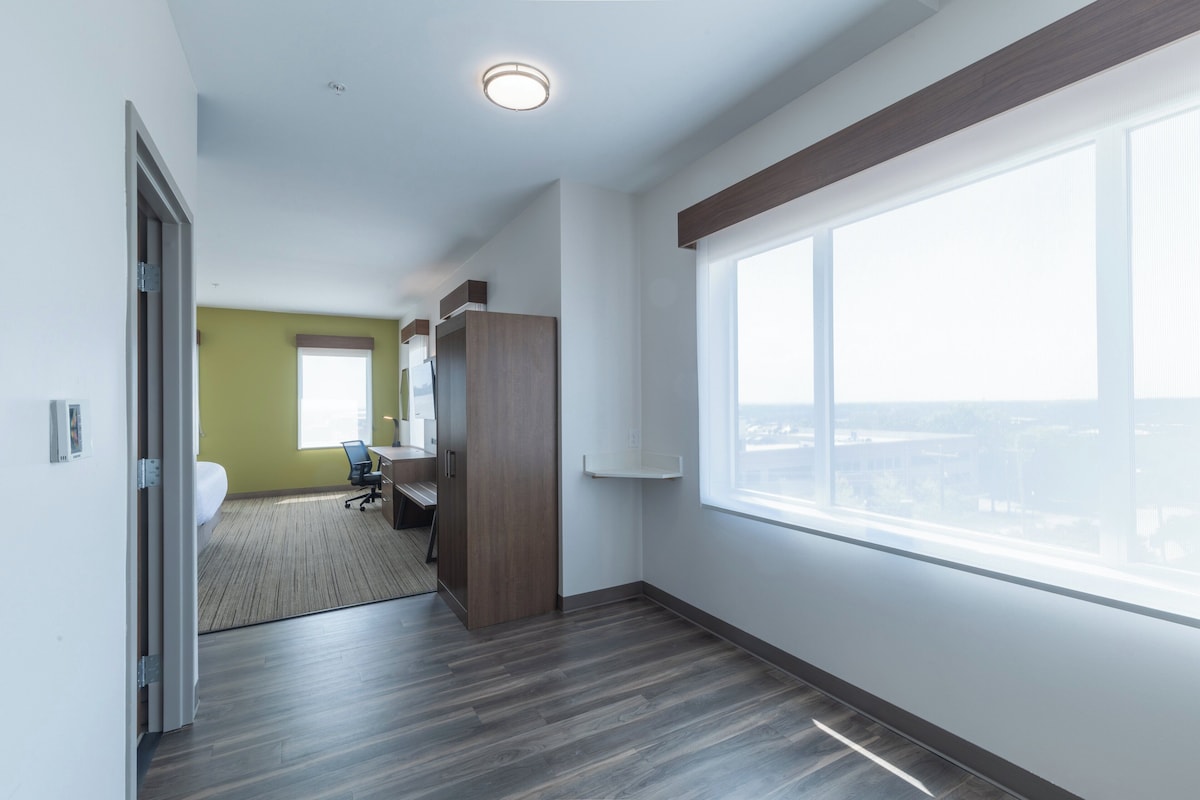 Une chambre d’hôtel avec une grande fenêtre, un bureau, une chaise et une armoire.