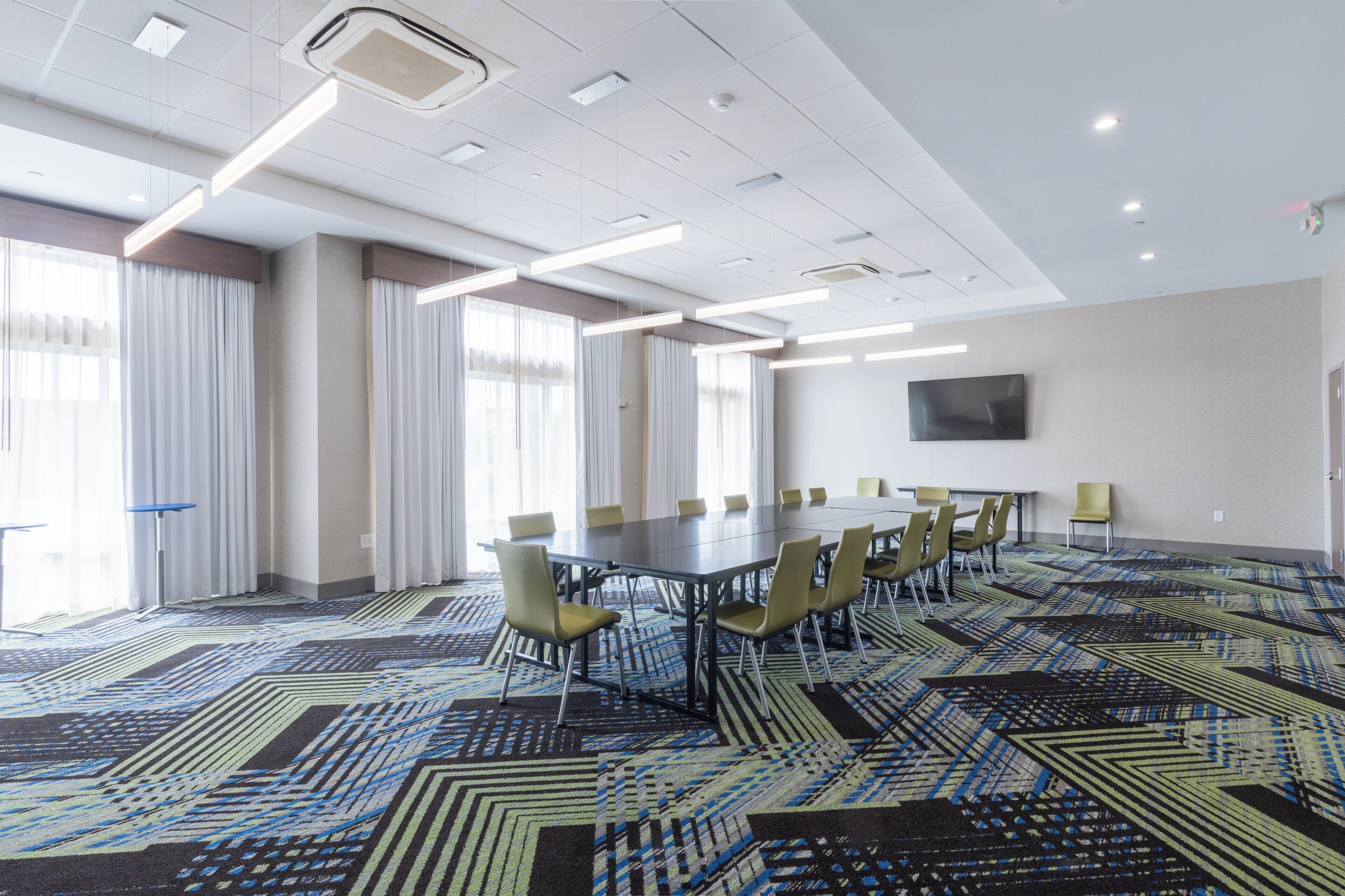 A conference room with a large table, chairs, a flat-screen TV on the wall, and patterned carpet.