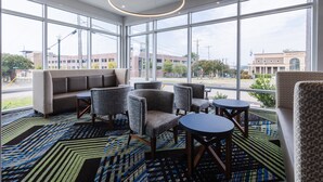 A modern hotel lobby with large windows, a patterned carpet, and a seating area with sofas and chairs.