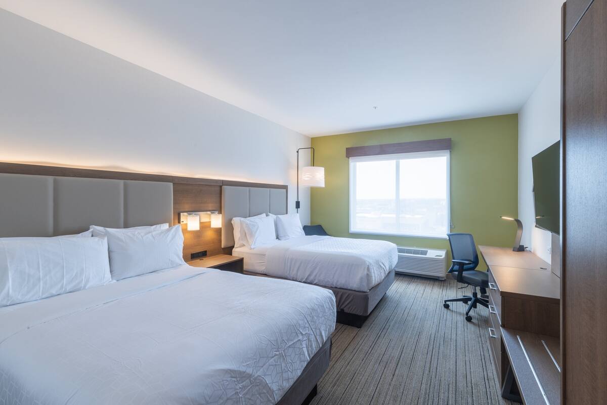 A hotel room with two beds, a desk, a chair, a lamp, and a large window.