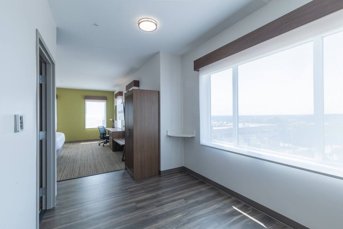A hotel room with a large window, a desk, a chair, and a wardrobe.