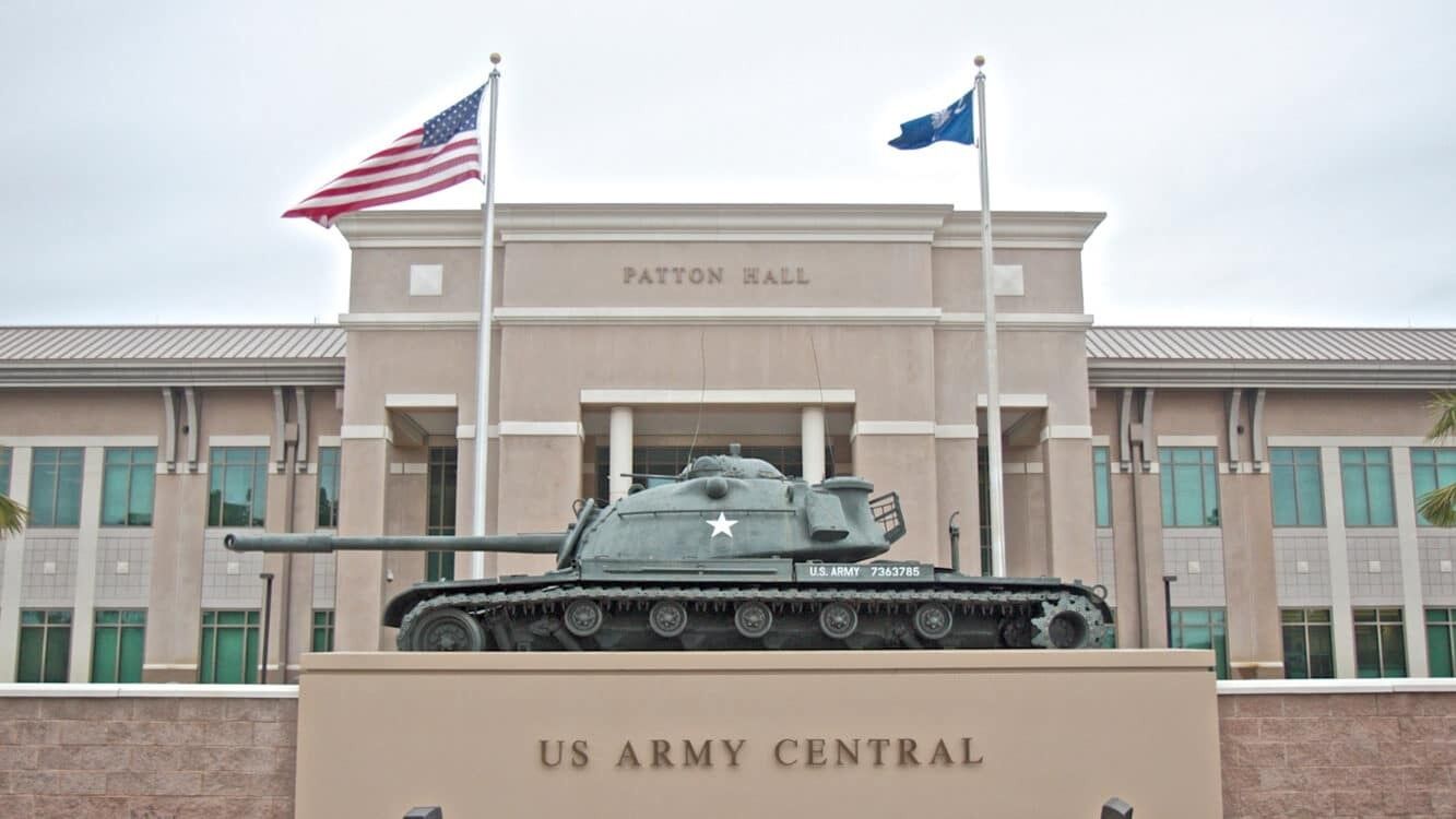 A US Army Central building with a tank in front, two flags, and a clear sky.