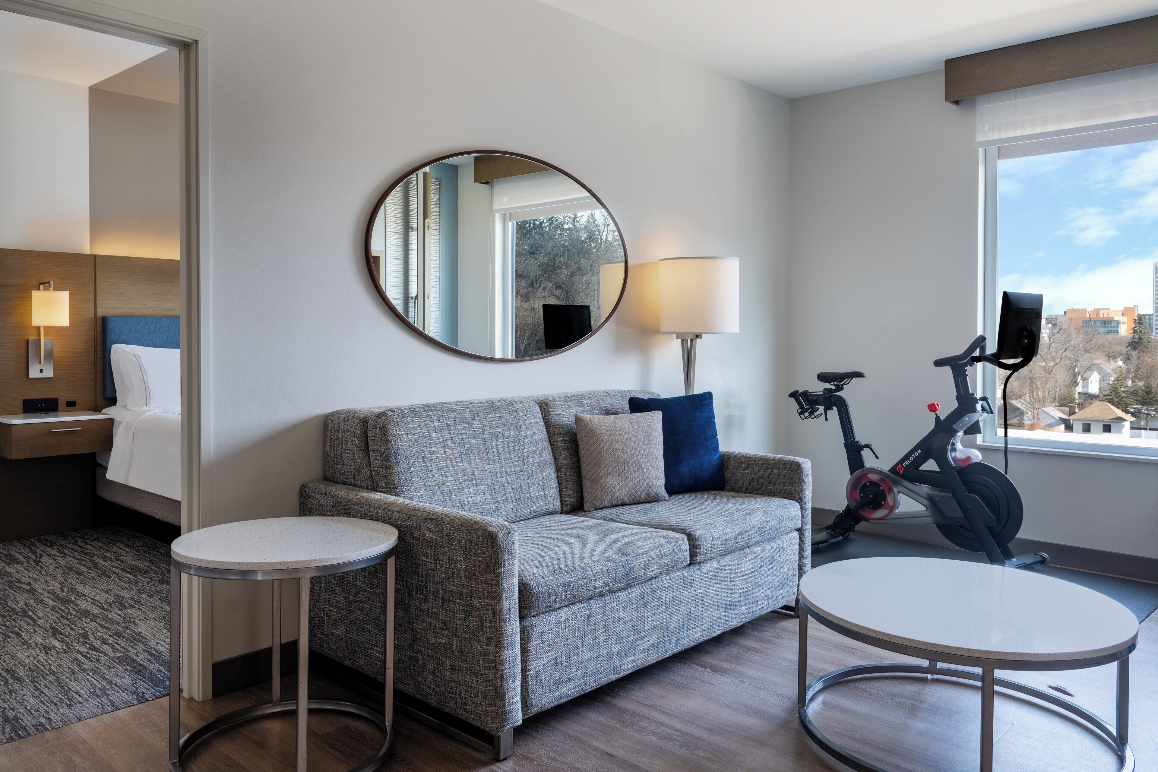 A hotel room with a grey sofa, a round mirror, a small round table, a standing lamp, and a stationary exercise bike.