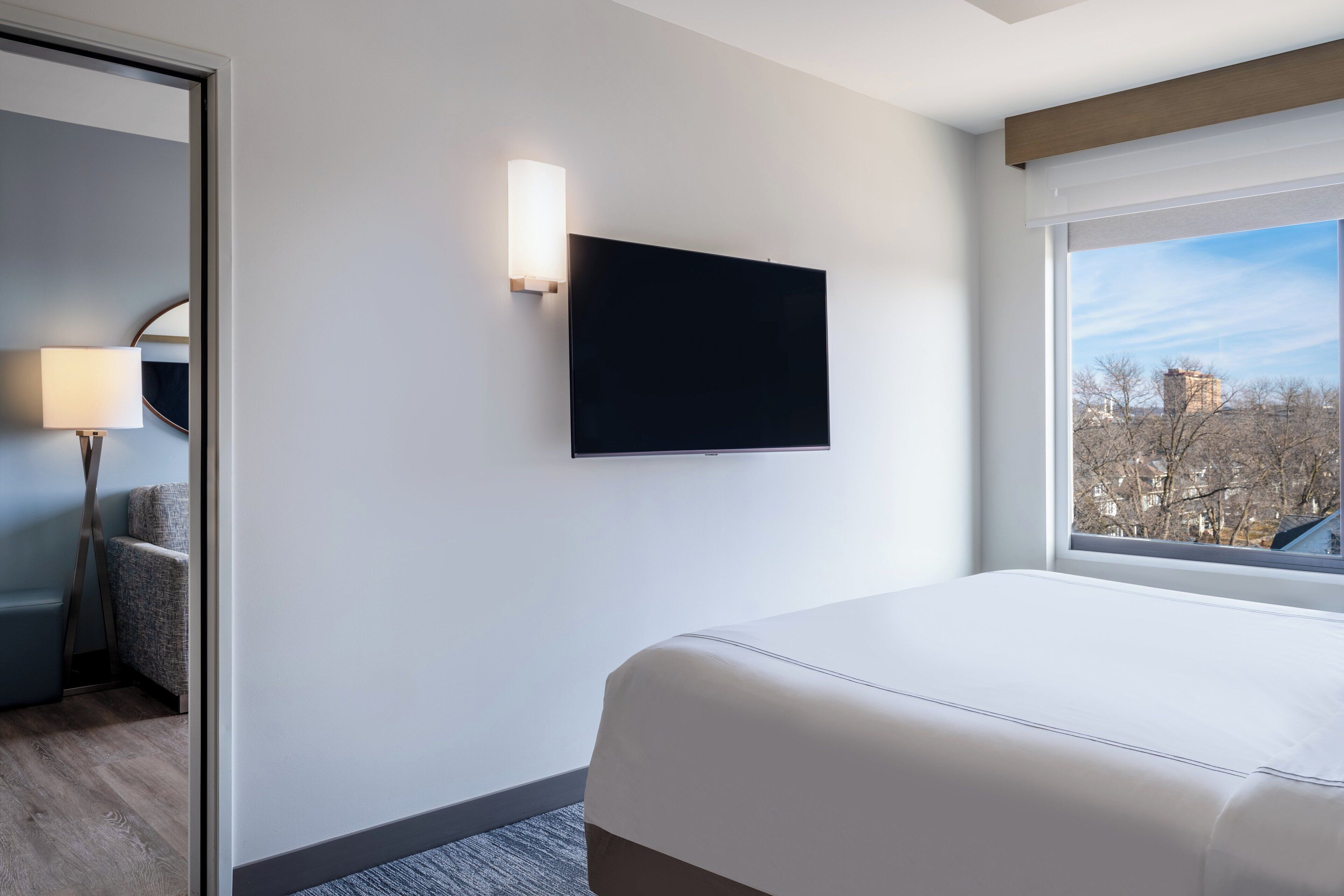 A hotel room with a bed, a TV mounted on the wall, a lamp, and a view of the city through a window.