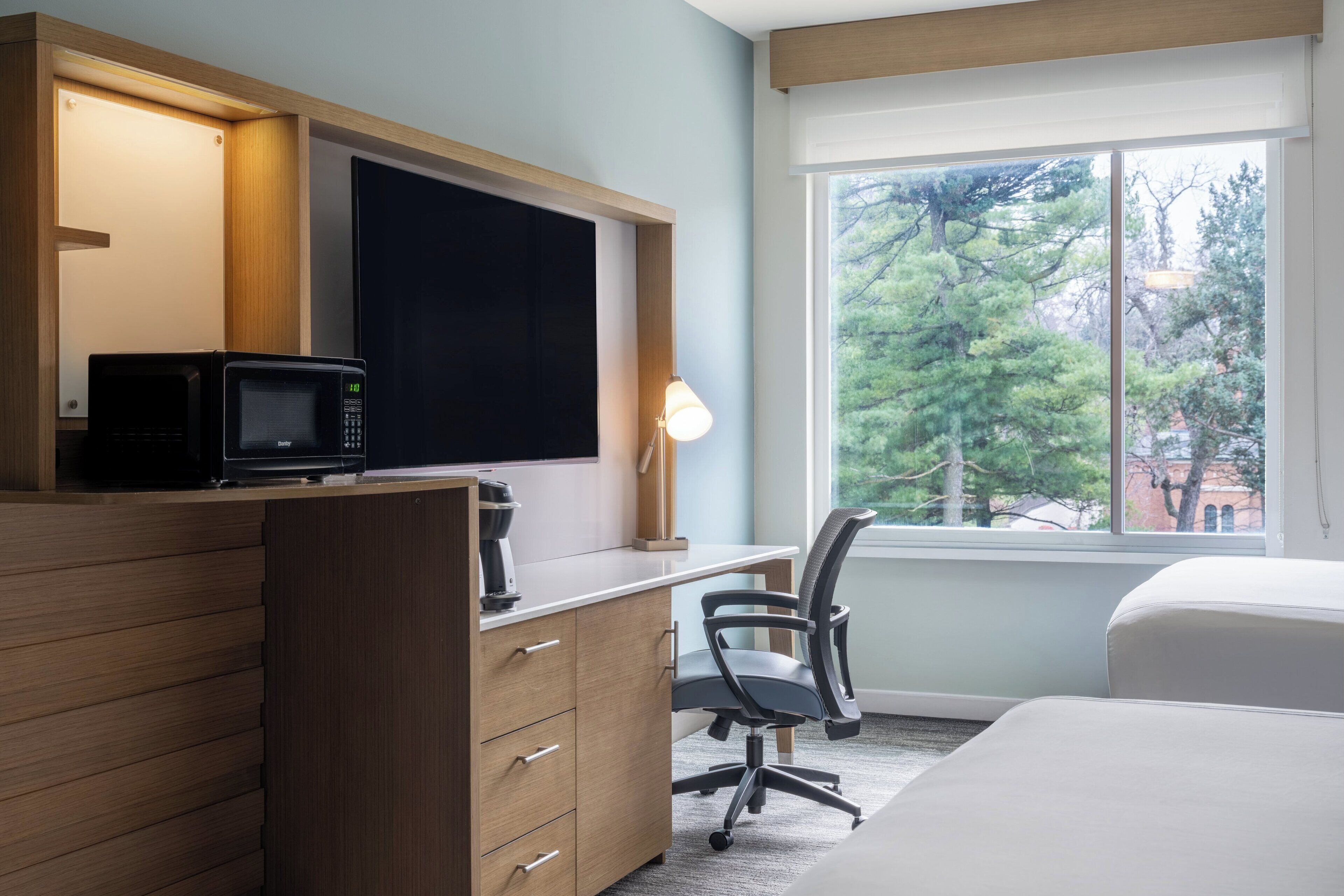 A hotel room with a wooden desk, a black microwave, a lamp, a chair, and a large window with a view of trees.