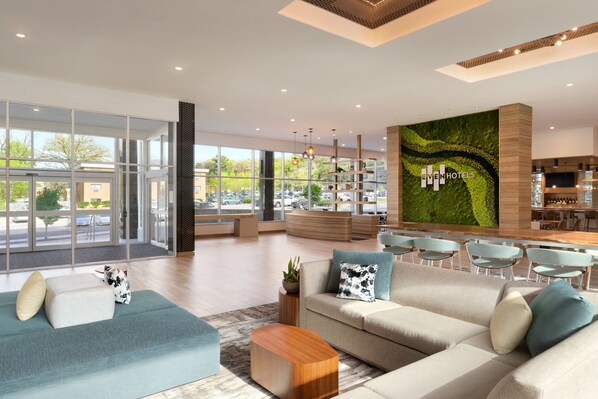 Un vestíbulo de hotel moderno con un gran muro de vidrio, un mural de pared verde y un área de descanso con sofás y sillones.