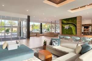 Un vestíbulo de hotel moderno con un gran muro de vidrio, un mural de pared verde y un área de descanso con sofás y sillones.