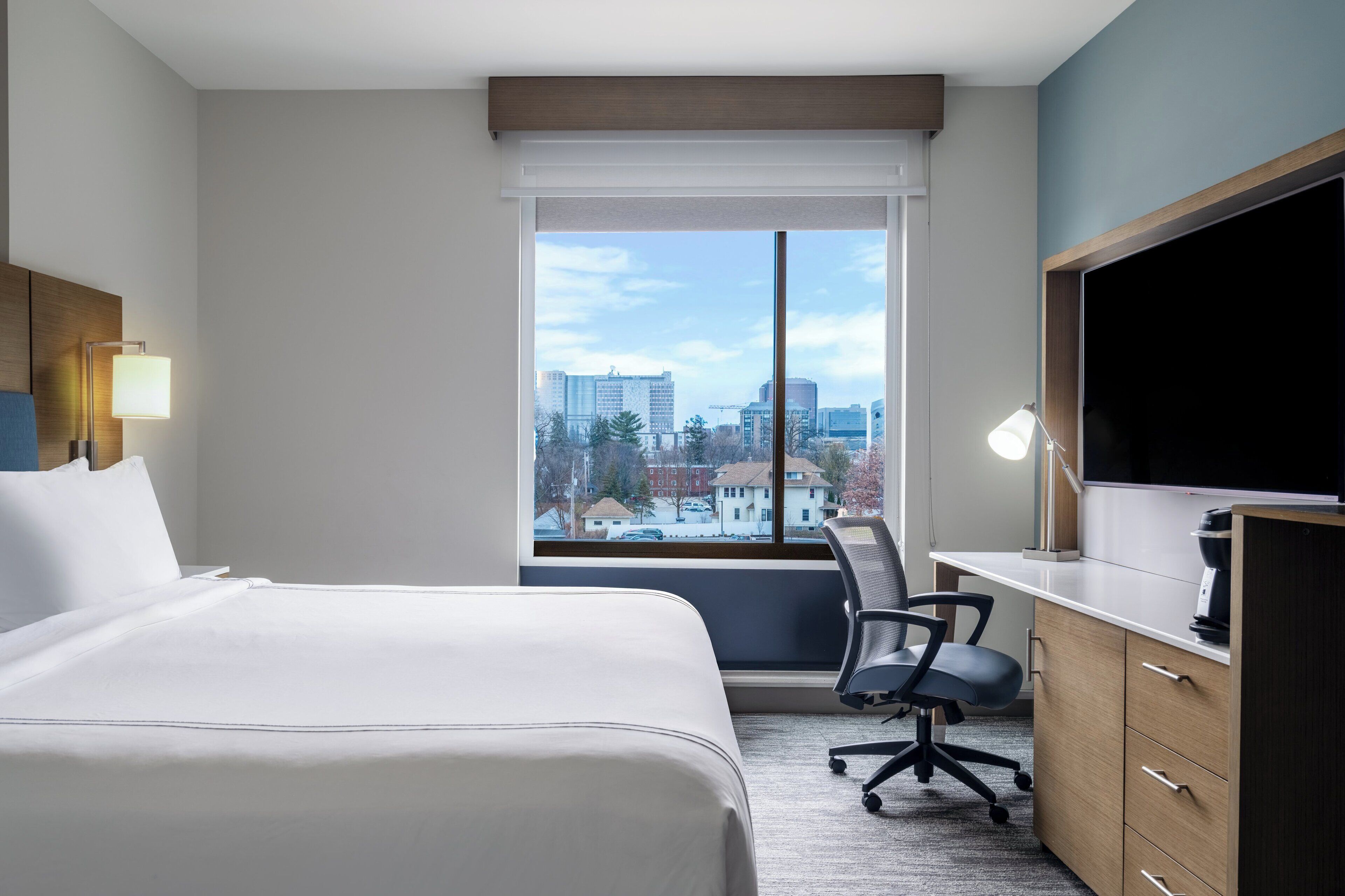 A hotel room with a bed, desk, chair, and a large window with a city view.